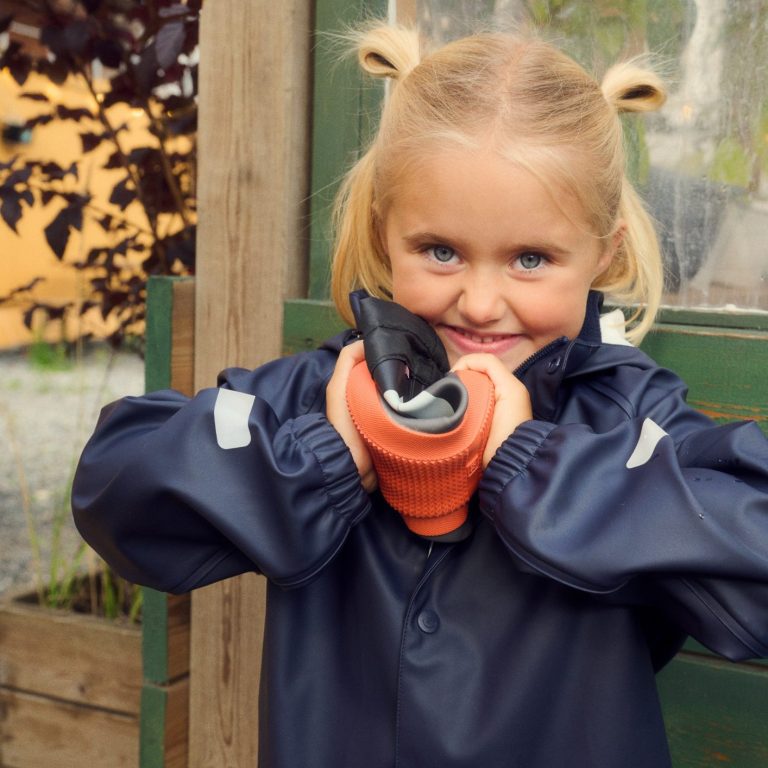 Ein lachendes Mädchen in blauer Jacke hält einen Gummistiefel in der Hand.